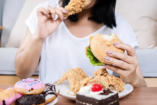 Mulher comendo um hambúrguer diante de um prato com doces e frituras