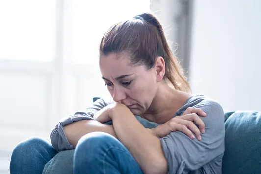 Mulher abraçando os joelhos no sofá, com expressão de tristeza